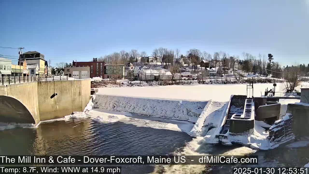 A partially frozen river flows past a dam and a snow-covered town under a clear blue sky.