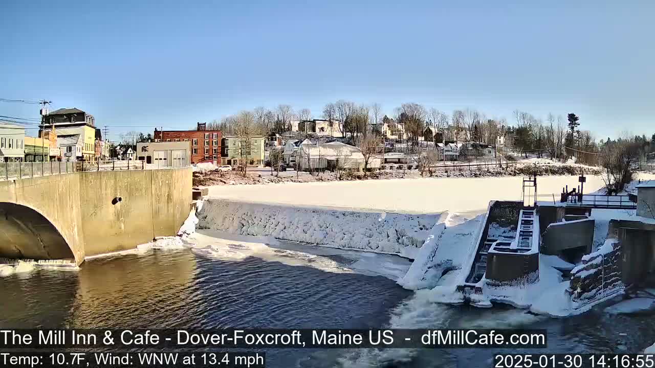 A partially frozen river flows under a bridge past a dam in a small town on a sunny, cold day.