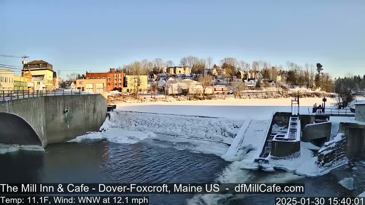 A snow-covered town is viewed from a bridge overlooking a partially frozen river with a dam and lock system.