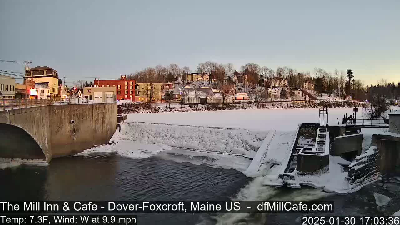 A snow-covered town is seen from a bridge overlooking a partially frozen river with a dam and lock system.
