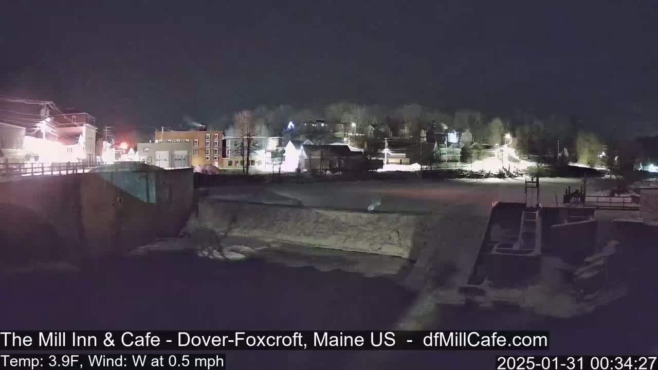 A nighttime view of a snow-covered town with a frozen river or canal in the foreground, under a dark sky with a temperature of 3.9°F and a west wind of 0.5 mph.