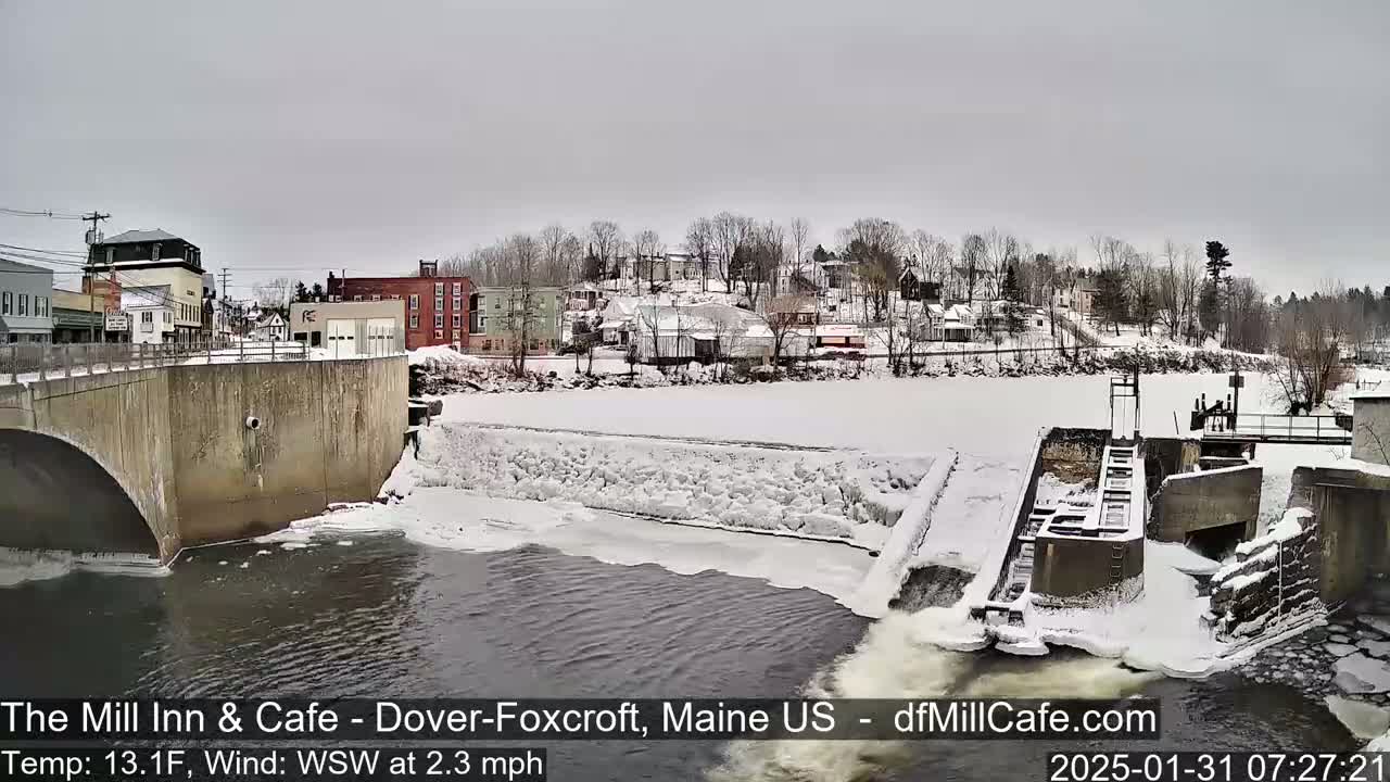 A snow-covered town overlooks a partially frozen river with a dam and flowing water.