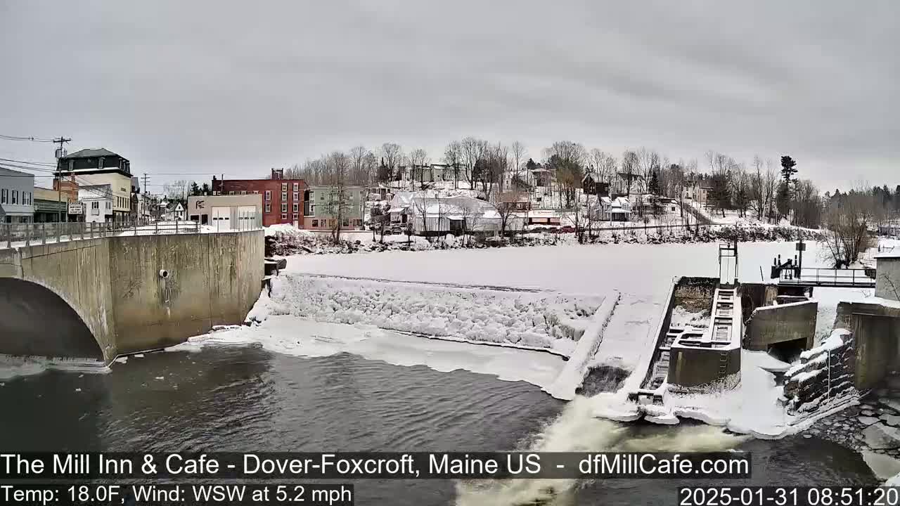 A snow-covered town is seen from a bridge overlooking a partially frozen river with a dam and flowing water.