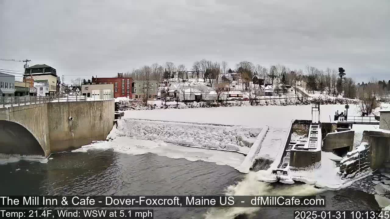 A snow-covered river flows past a dam and a town under a cloudy sky.