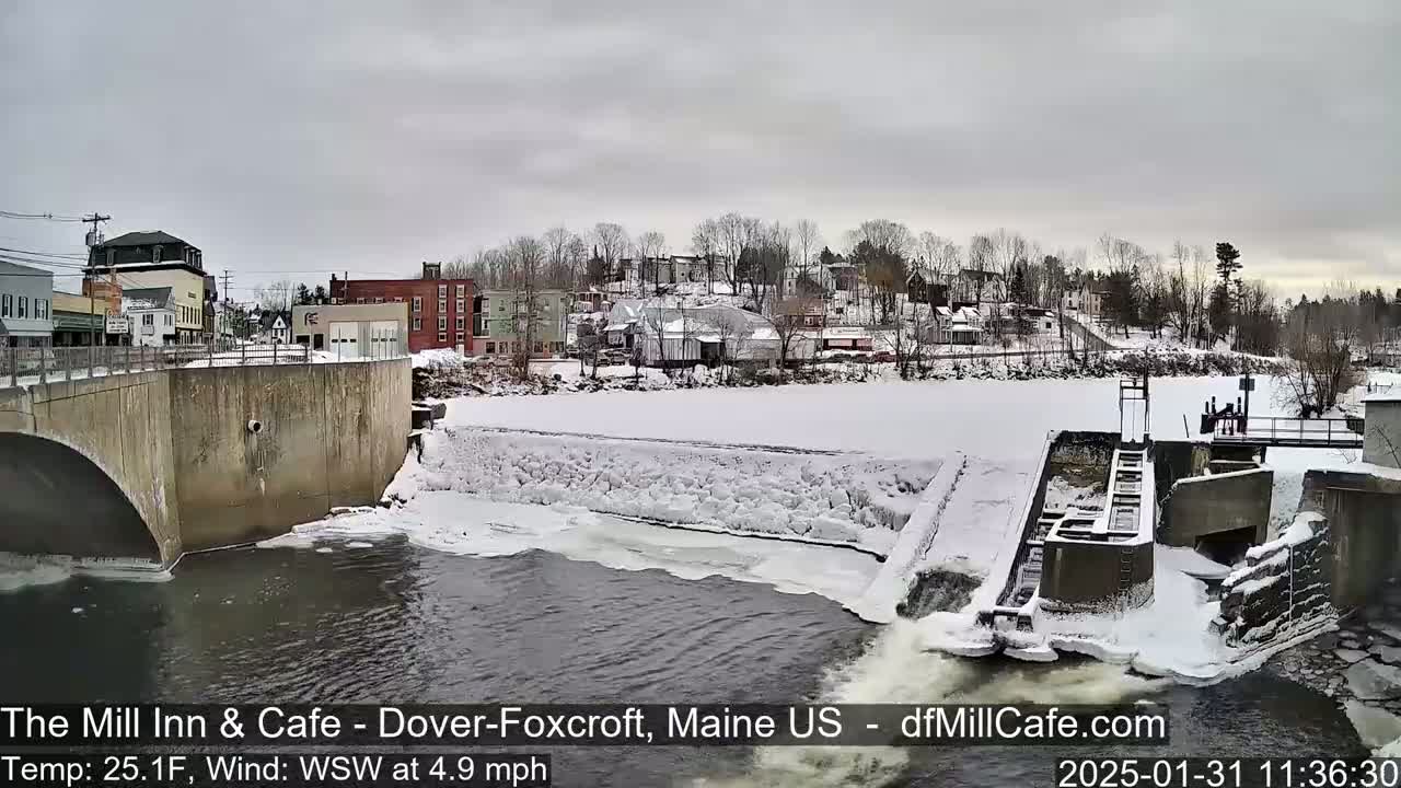 A snow-covered town is seen from a bridge overlooking a partially frozen river with a dam and flowing water.