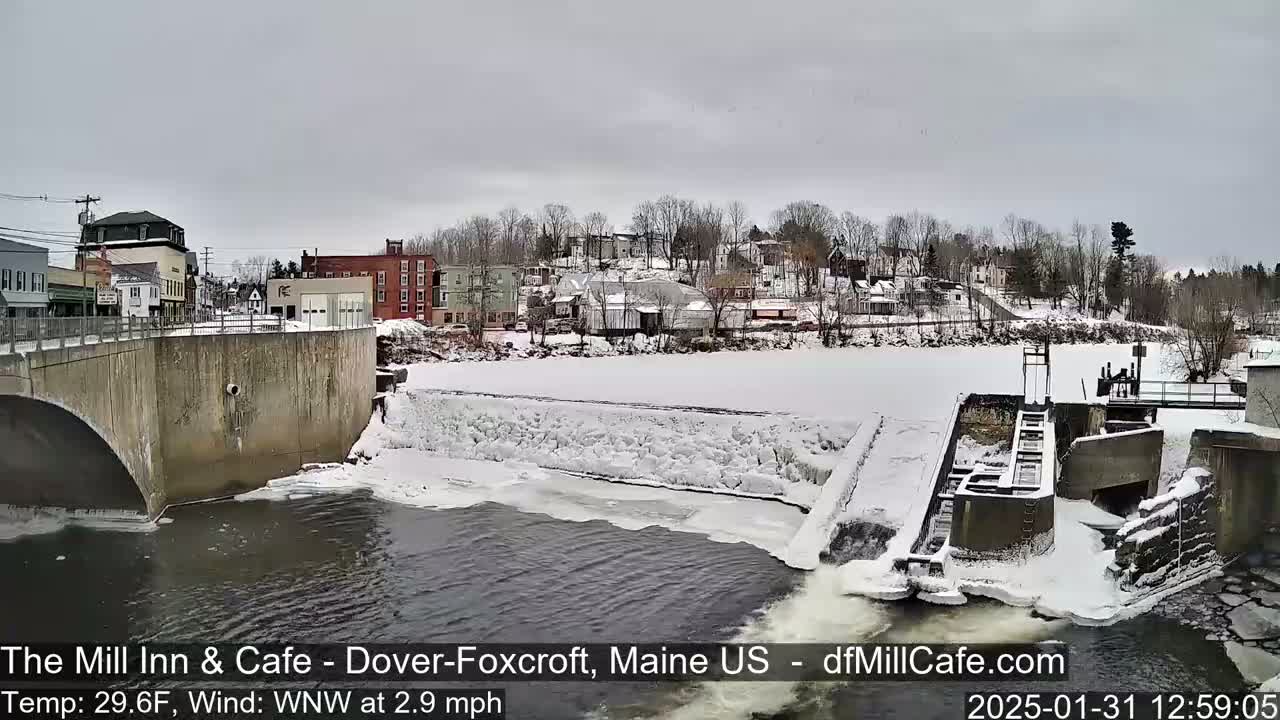 A snow-covered dam and river are seen below a bridge and a small town on an overcast day.