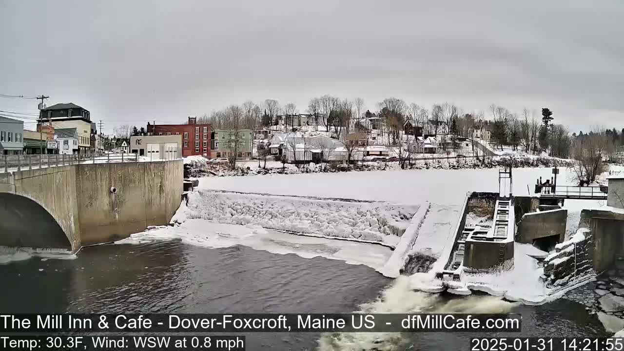 A snow-covered town sits beside a partially frozen river with a dam and flowing water, under an overcast sky.
