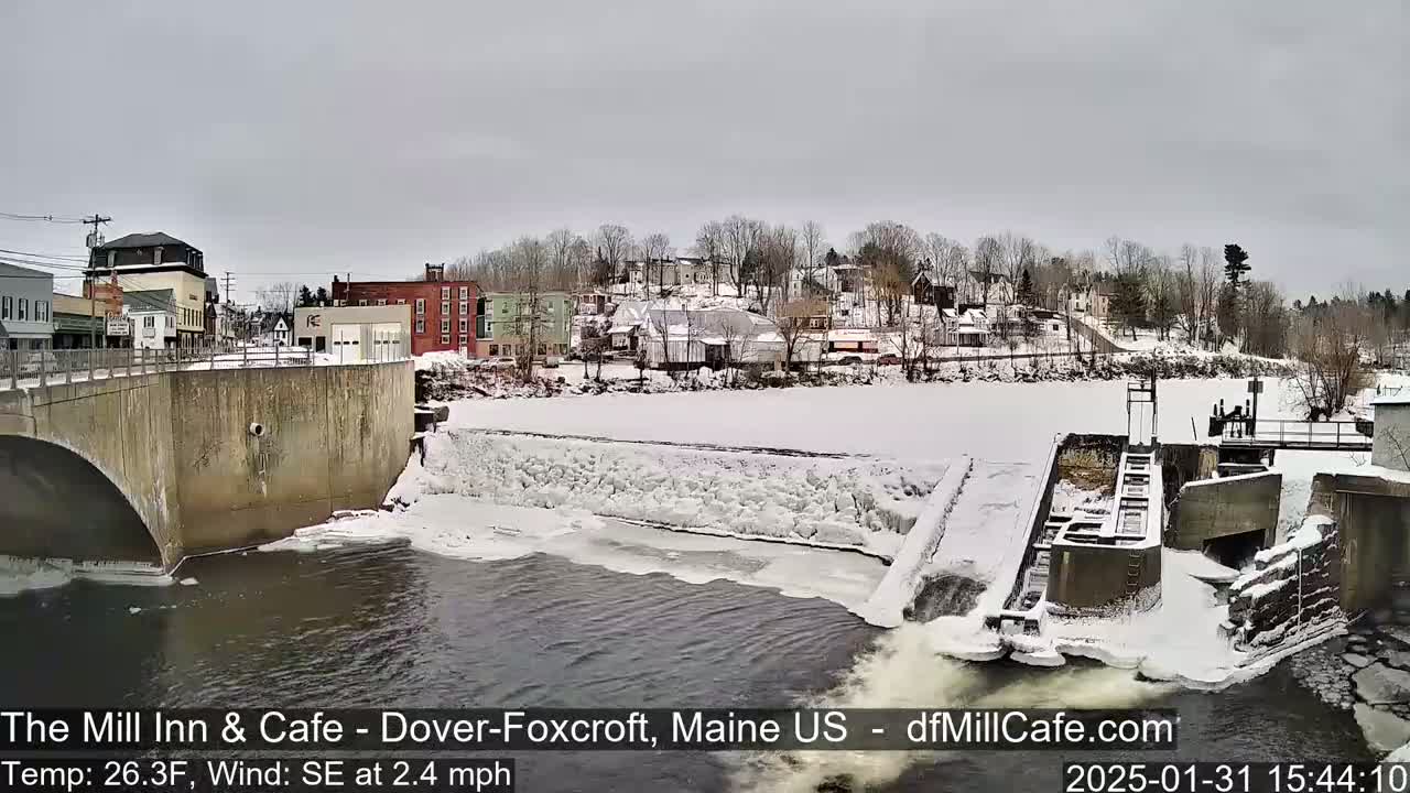 A snow-covered town is viewed from a bridge overlooking a partially frozen river with a dam and flowing water.
