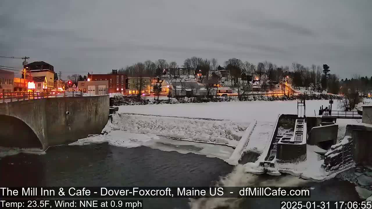 A snow-covered town sits beside a partially frozen river with a dam and lock system, under a twilight sky.