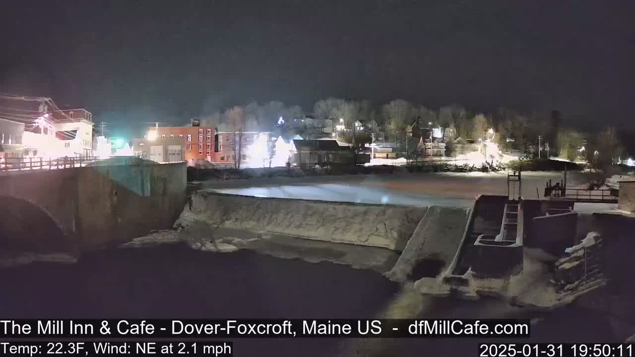 A nighttime view of a snow-covered town with a partially frozen river and dam under a clear, dark sky.
