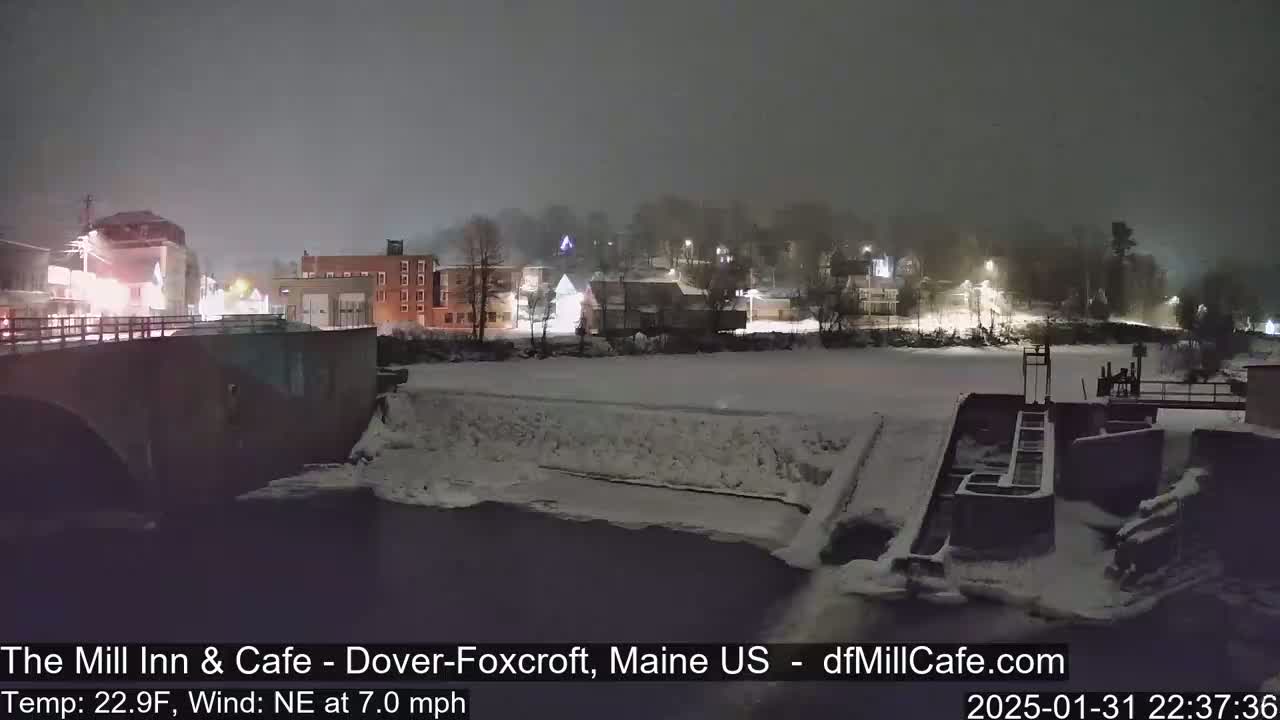 A snowy, nighttime scene shows a town with buildings and a bridge over a river, which is partially frozen and has a dam or lock system.