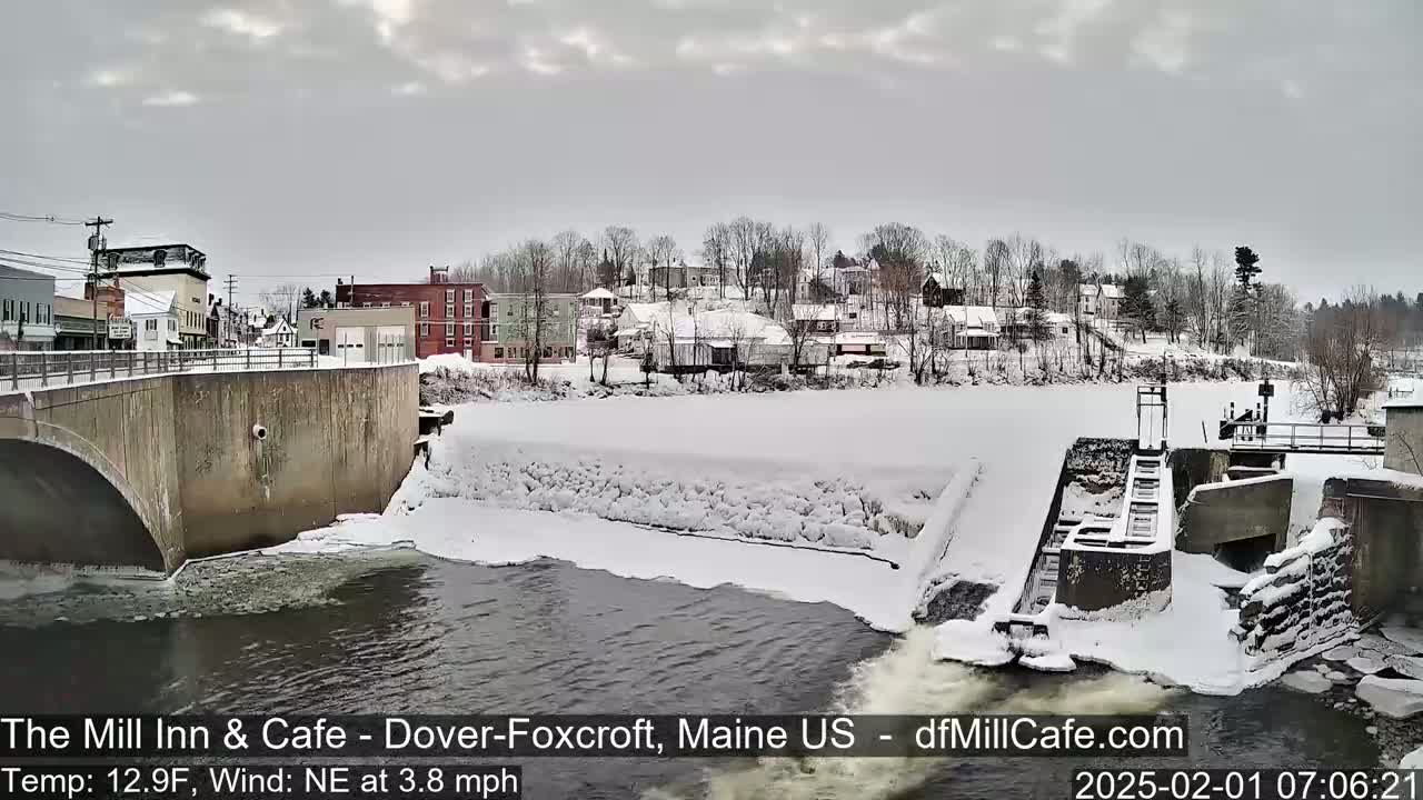 A snow-covered town with buildings and a dam on a river under a cloudy sky.
