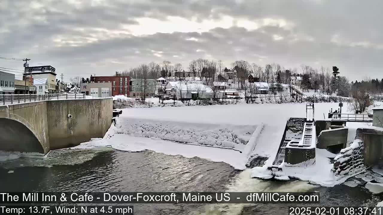 A snow-covered town, viewed from a bridge over a partially frozen river with a dam and a low water flow under an overcast sky.