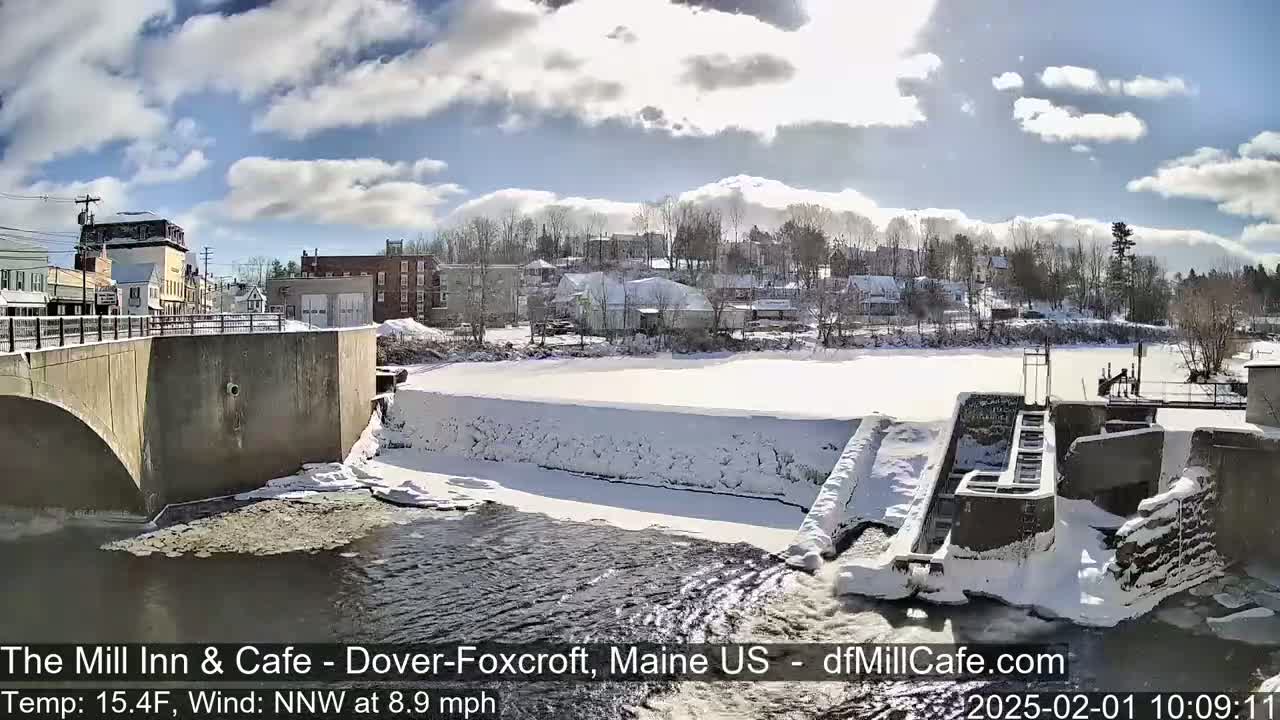 A snow-covered town and dam are visible under a partly sunny sky, with a river flowing past.