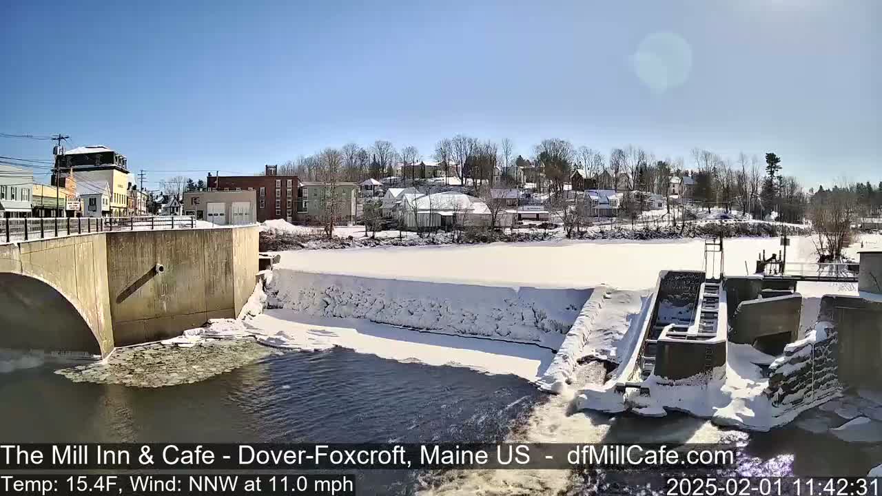 A snow-covered dam and river are seen from a bridge overlooking a small town on a sunny winter day.