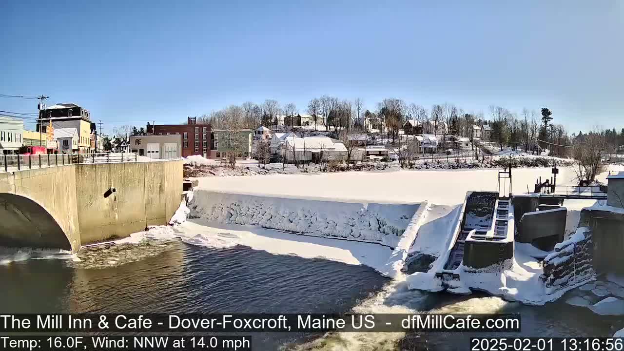 A snow-covered dam and river are viewed from a bridge, with a small town visible in the background under a clear, sunny sky.