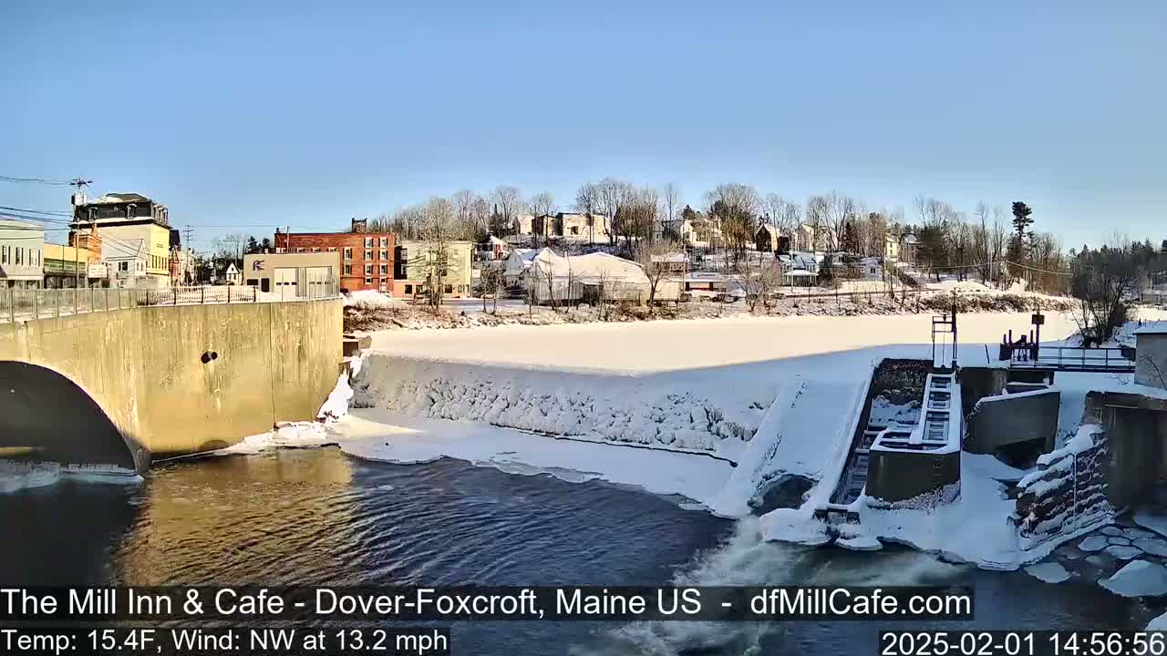 A snow-covered dam and river flow past a small town under a clear blue sky.