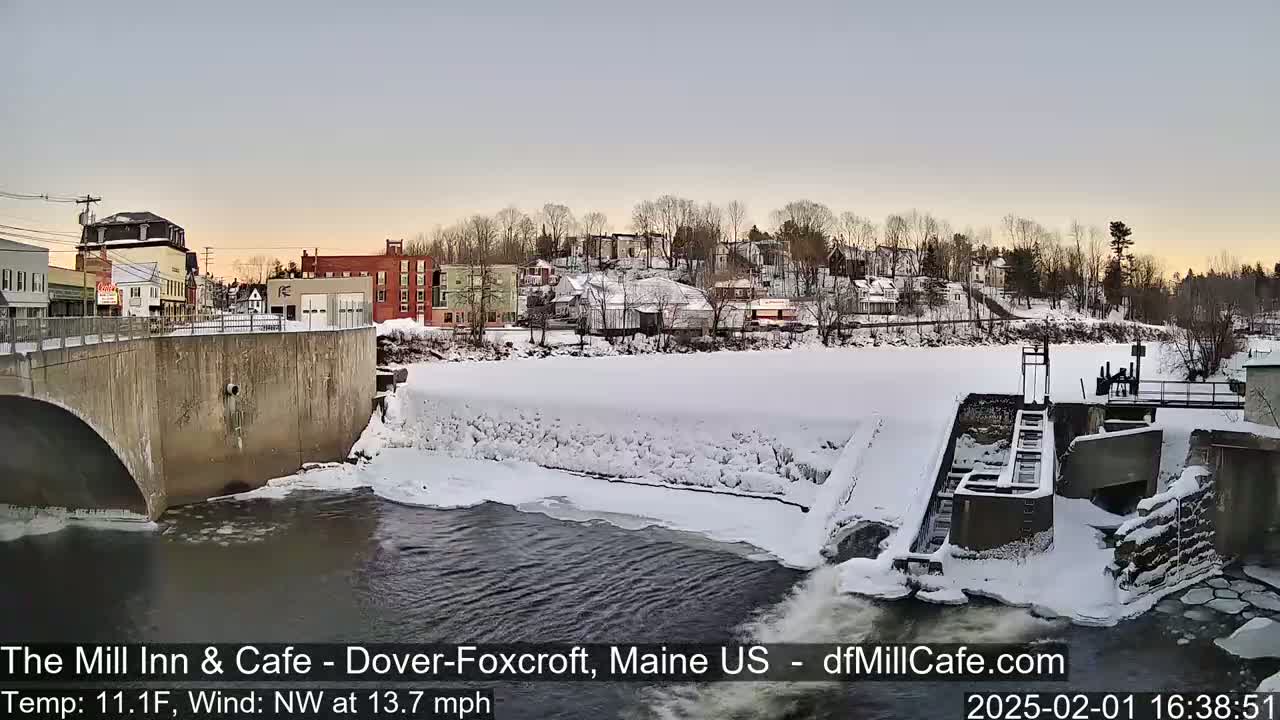 A snow-covered town and river are seen from a bridge, with a dam and partially frozen river in the foreground under a twilight sky.