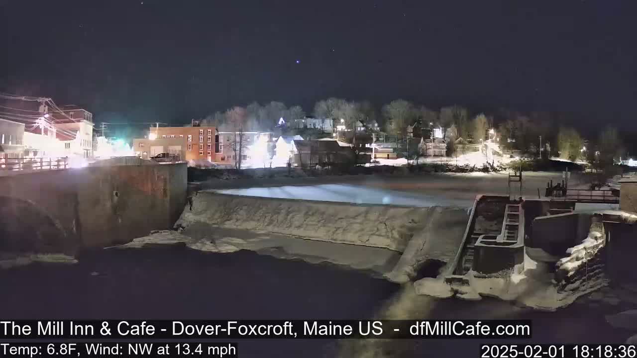 A nighttime view of a snow-covered dam and river, with a small town illuminated in the background under a clear, starry sky.