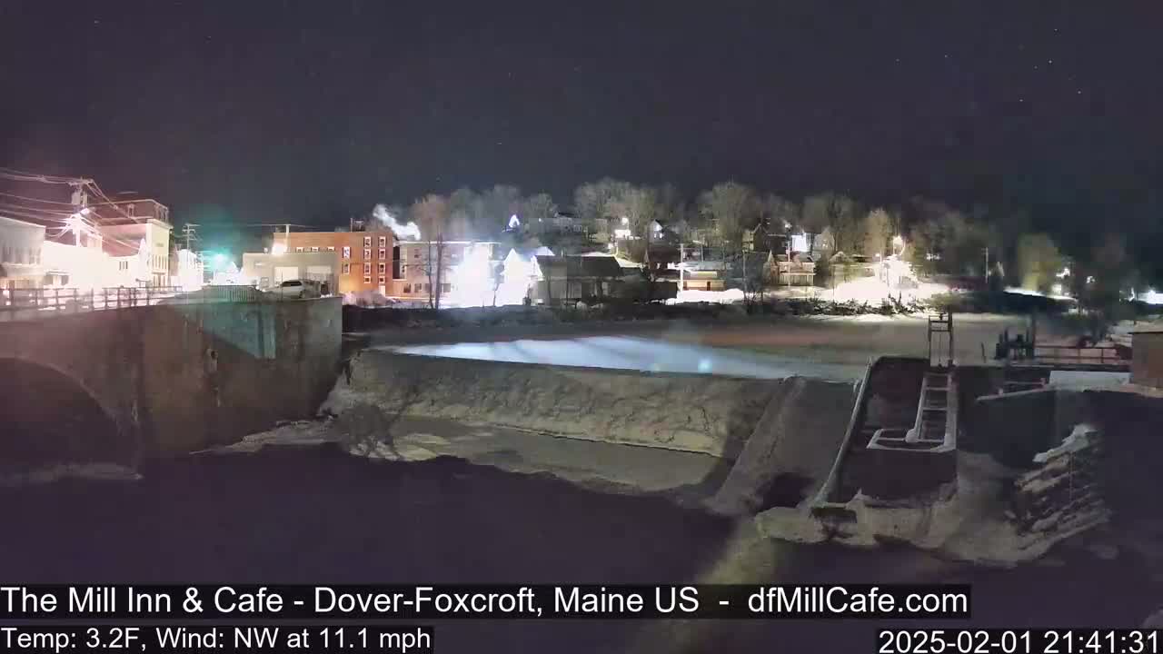 A nighttime view of a town's illuminated buildings and a river dam with a northwest wind at 11.1 mph and a temperature of 3.2 degrees Fahrenheit.