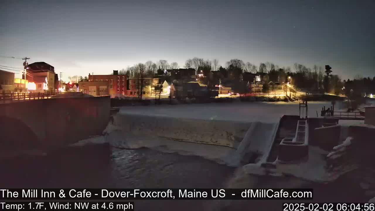 A nighttime view of a snow-covered river flowing past a dam and a small town with illuminated buildings under a clear, dark sky.