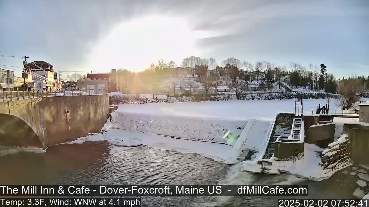 A partially frozen river flows past a dam and snow-covered town under a partly sunny sky.