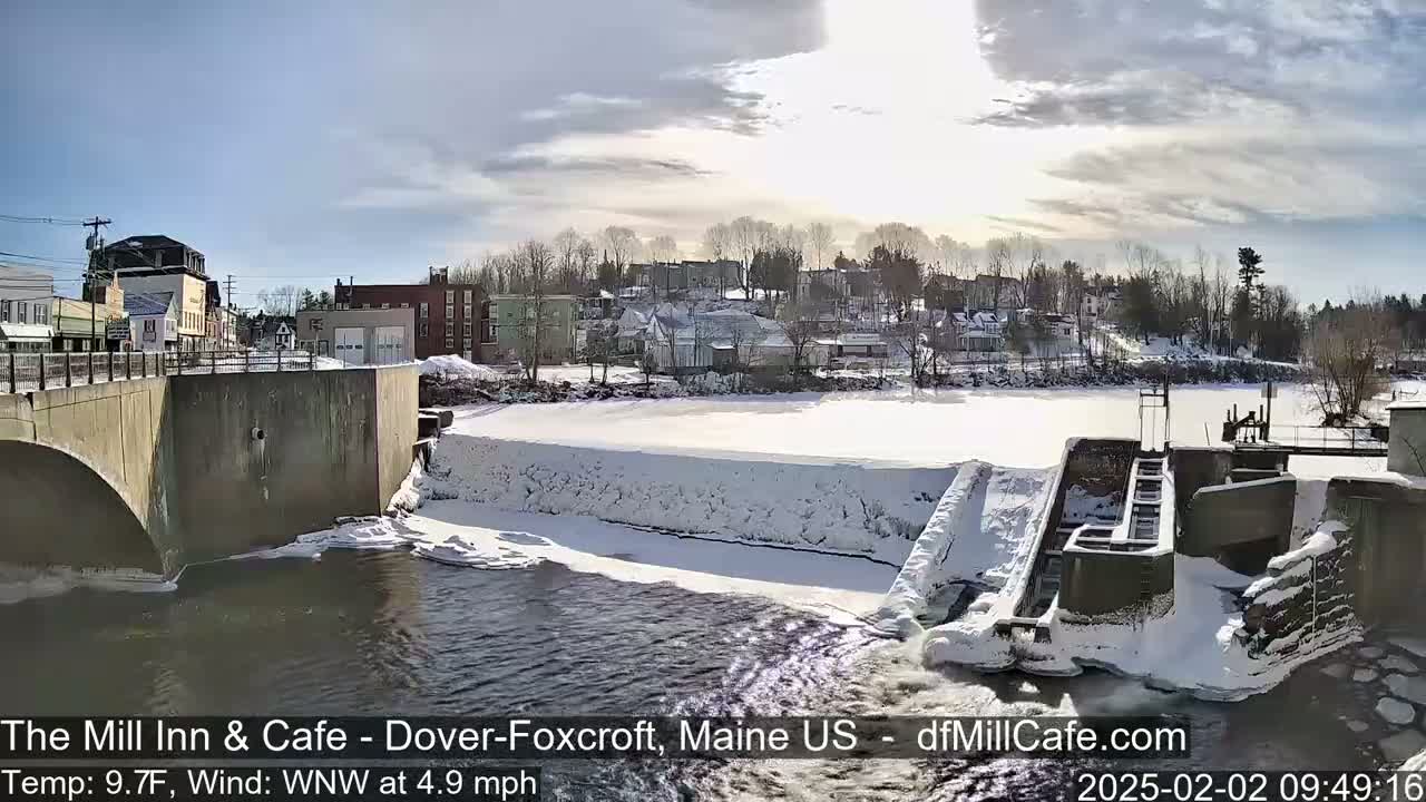 A snow-covered dam and river flow past a small town under a partly sunny sky.
