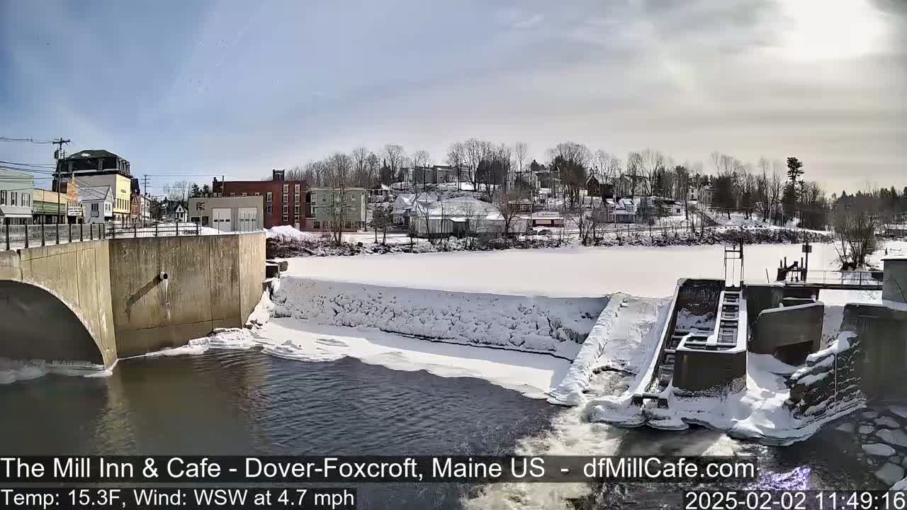 A snow-covered dam and river are seen below a bridge, with a small town in the background under a sunny sky.