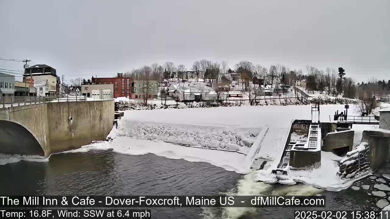 A snow-covered town with a partially frozen river and dam is seen from above a bridge on an overcast day.