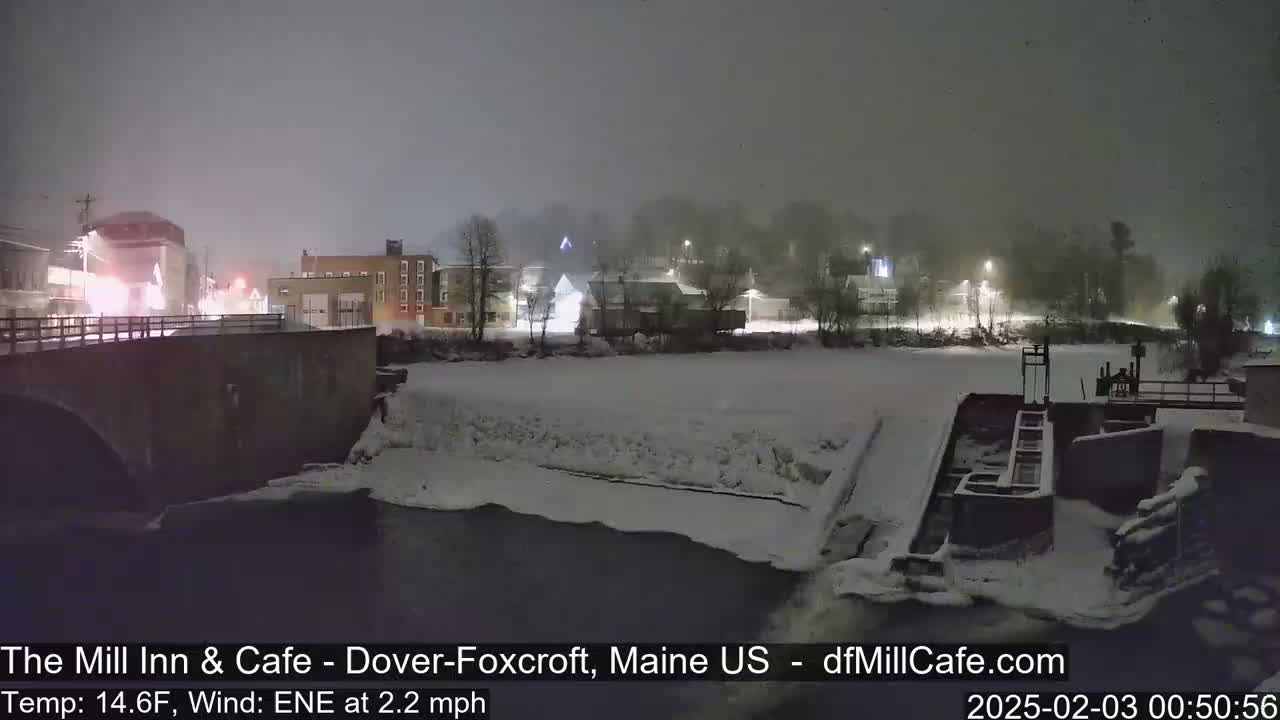 A snow-covered town at night is seen from a bridge overlooking a partially frozen river with a lock.
