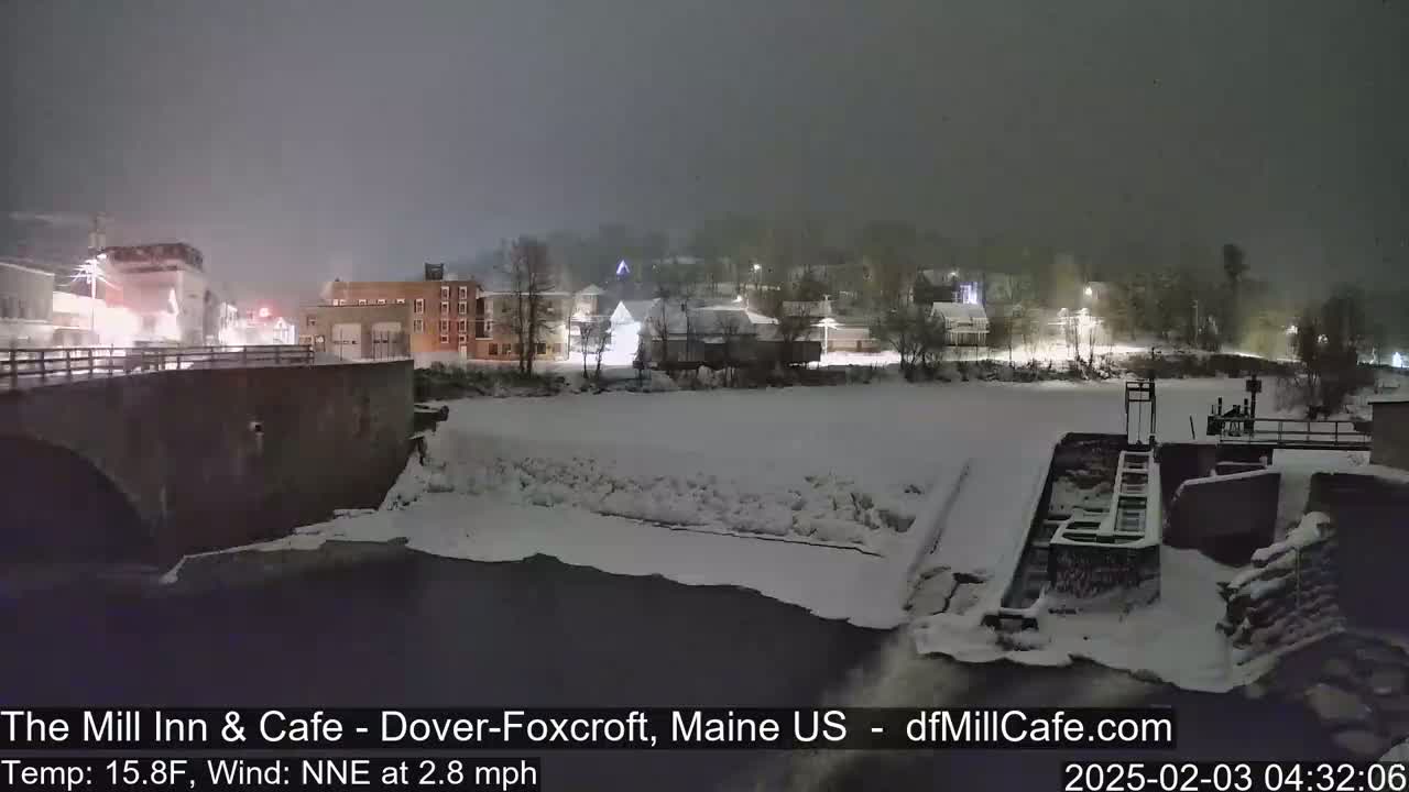 A snow-covered town at night is viewed from a bridge over a partially frozen river with a dam visible in the foreground, under falling snow.