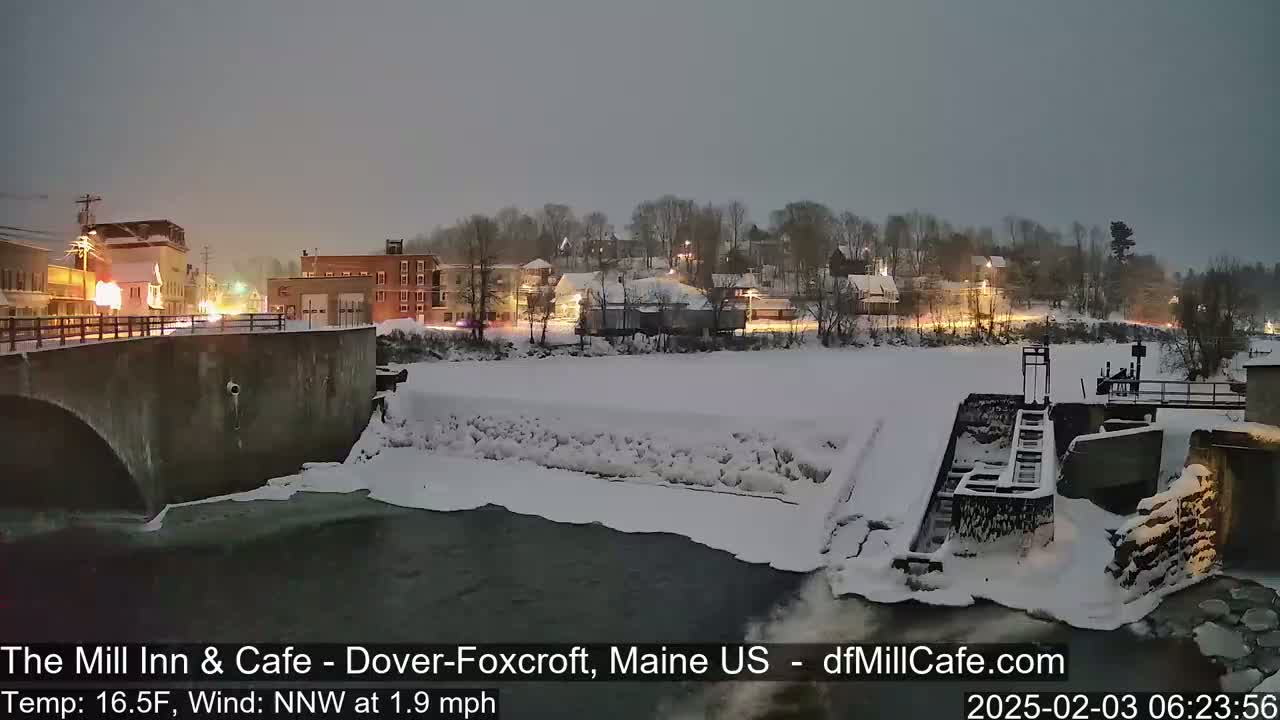 A snow-covered town at night is viewed from a bridge over a partially frozen river with a dam and lock visible in the foreground.
