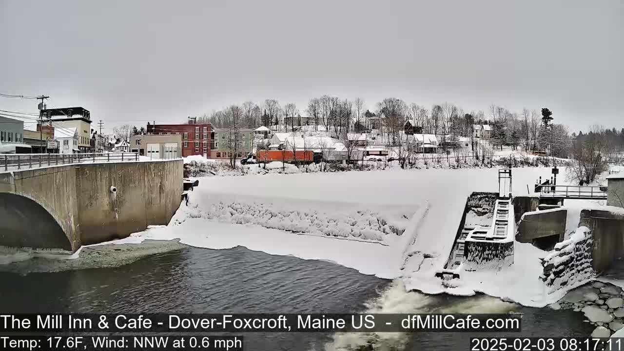 A snow-covered dam and river flow past a small town under a gray sky.
