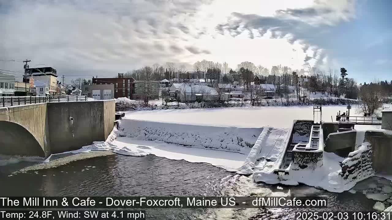 A snow-covered dam and town are seen under a partly sunny sky with a light southwest wind.
