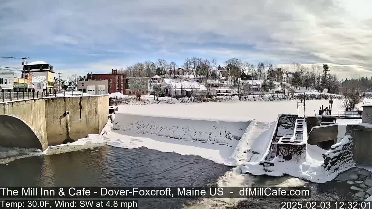 A snow-covered town is viewed from a bridge overlooking a dam and river on a partly sunny day.