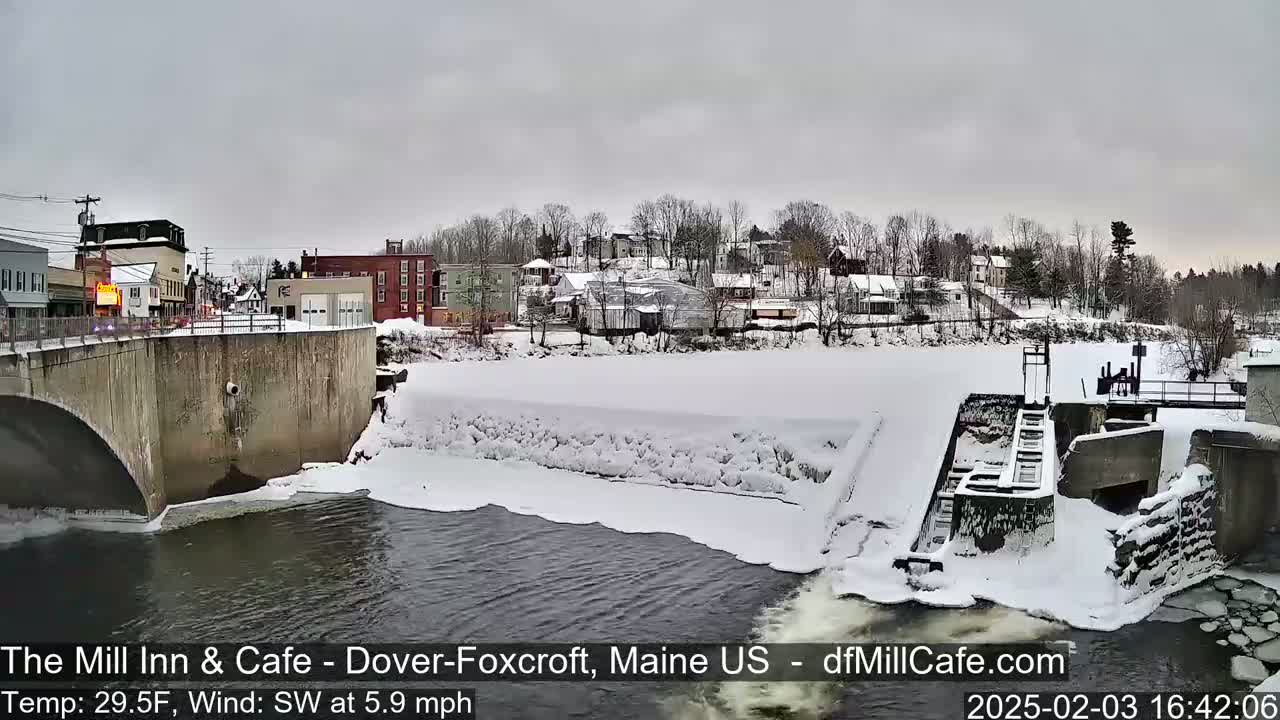 A snow-covered dam and river are seen from a bridge overlooking a small town under an overcast sky.