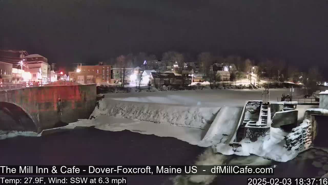 A snow-covered dam and river are illuminated at night by the lights of a nearby town.