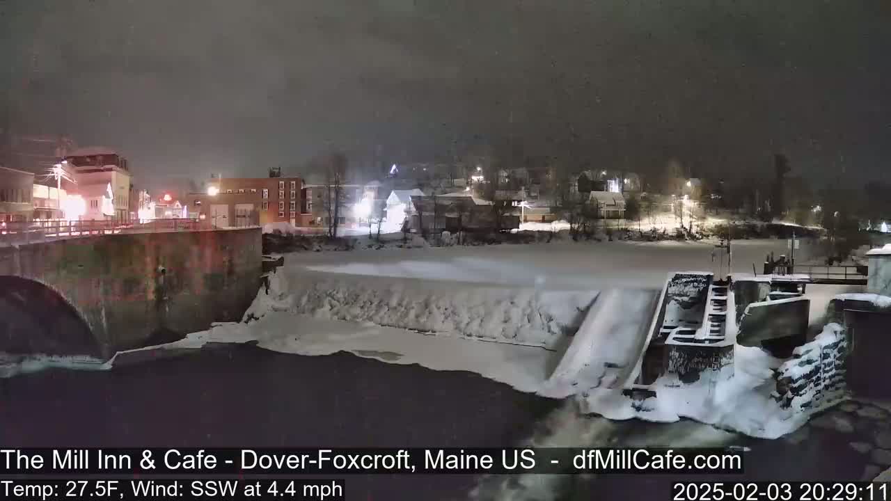 A snow-covered dam and partially frozen river are seen at night, under a lightly snowing sky, with a town in the background.