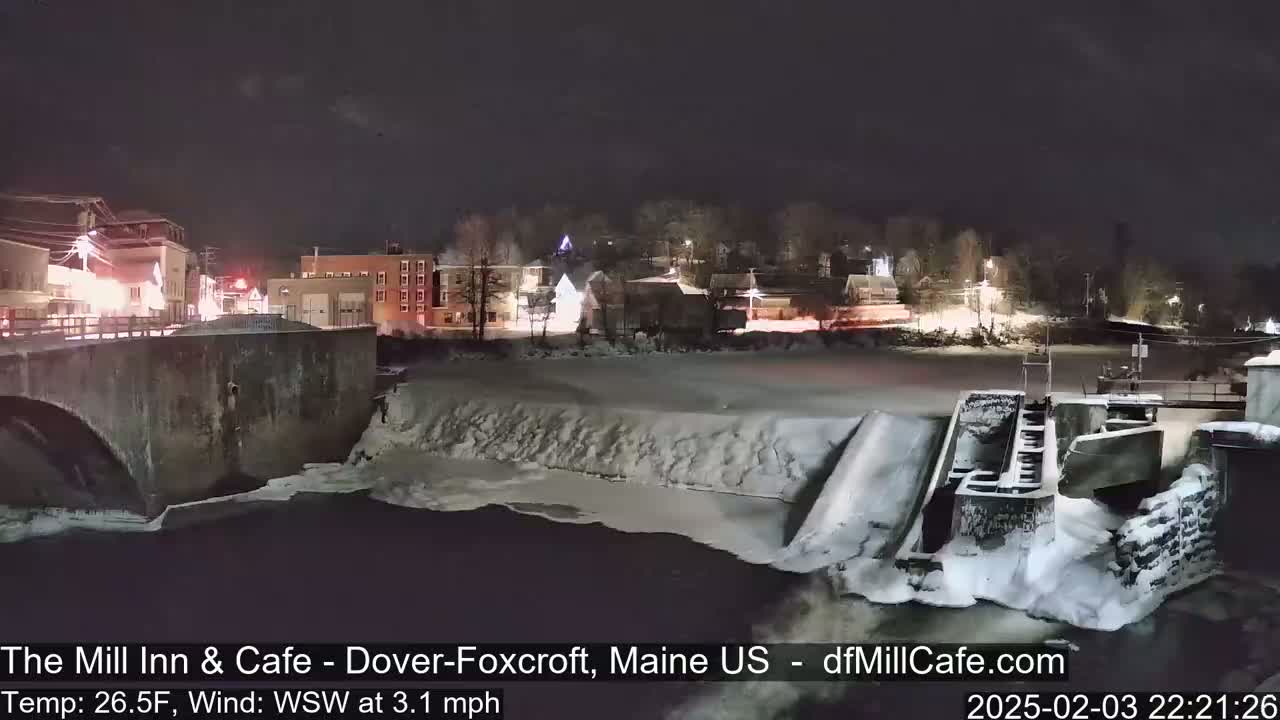 A snow-covered dam and river are illuminated at night by building lights, under a clear, dark sky.