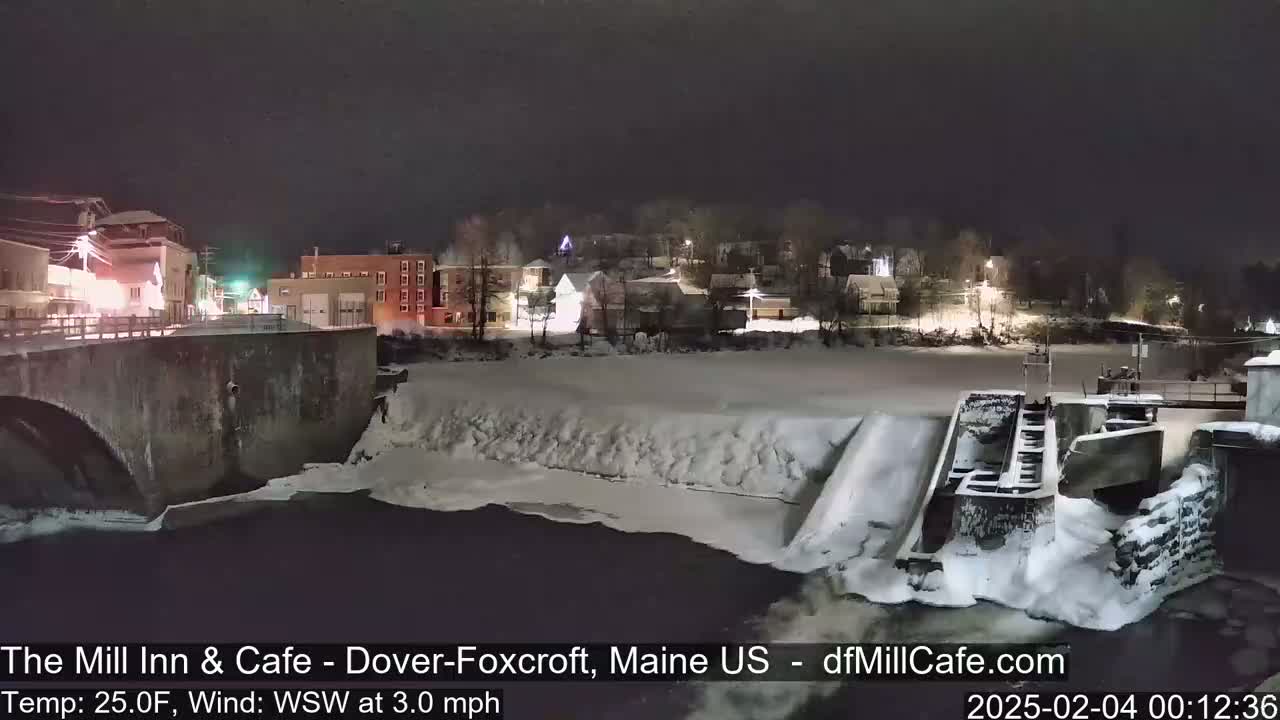A nighttime view of a snow-covered town and river with a dam, under a dark sky.