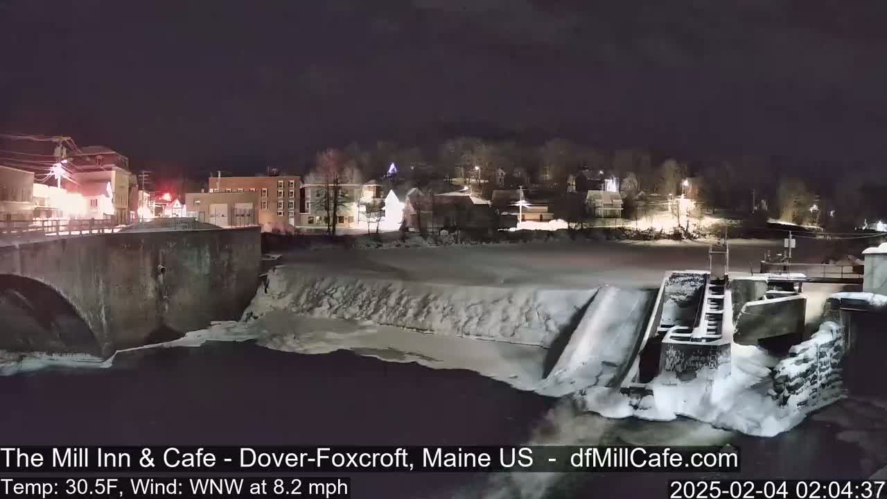 A snow-covered river flows beneath a bridge at night, past a dam and a small town with illuminated buildings.