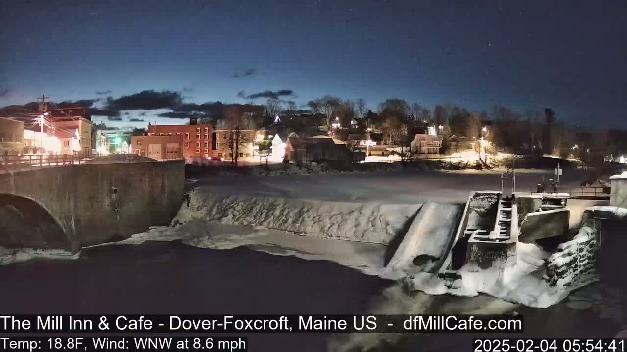 A nighttime view of a partially frozen river flowing past a dam and a town under a clear, dark sky.