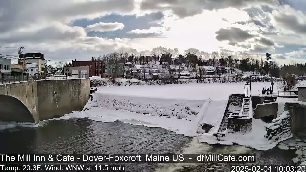 A snow-covered town is seen from a bridge overlooking a partially frozen river with a dam and a windy, 20.3°F day.