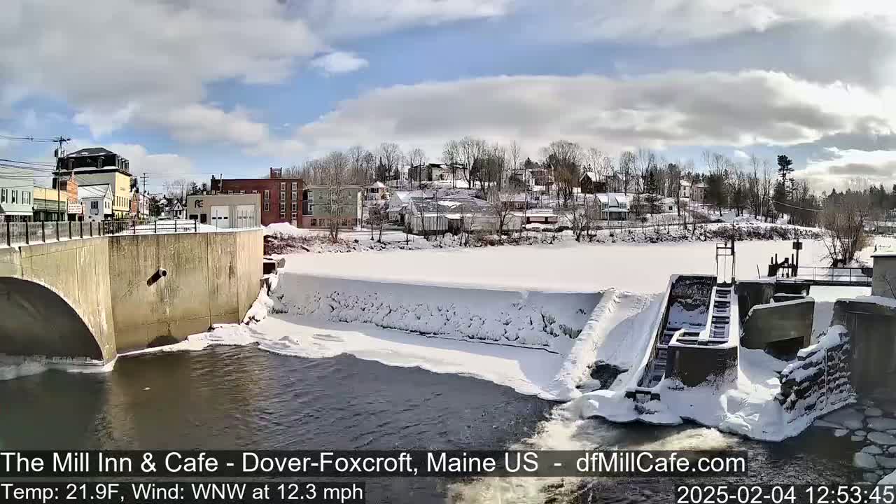 A snow-covered dam and river are seen from a bridge, with buildings and trees visible in the background under a partly cloudy sky.
