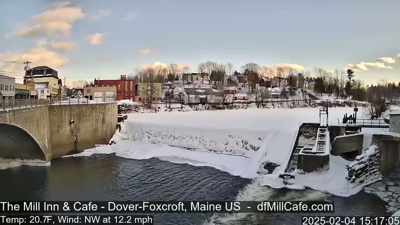 A snow-covered town is seen from a bridge overlooking a partially frozen river with a dam and a water lock.