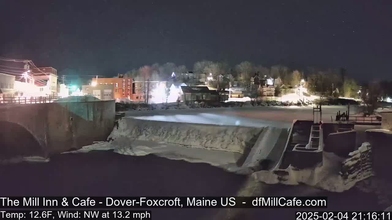 A nighttime view of a snow-covered dam and river flowing past a small town under a clear, dark sky.