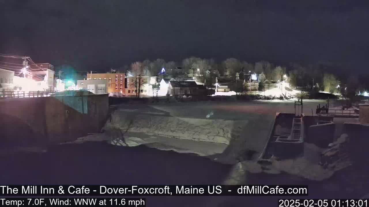 A nighttime view of a snow-covered town beside a partially frozen river, under a clear, dark sky with a wind blowing from the west-northwest at 11.6 mph and a temperature of 7°F.