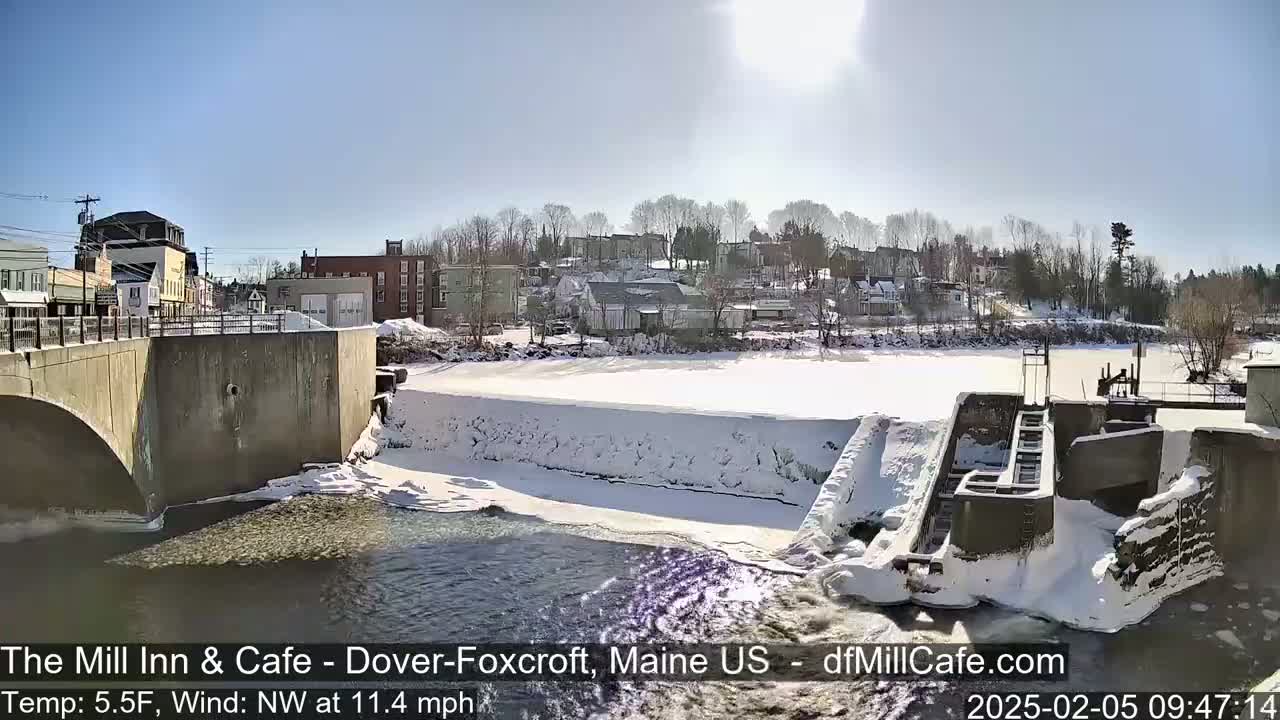 A snow-covered dam and river flow past a town under a sunny sky.