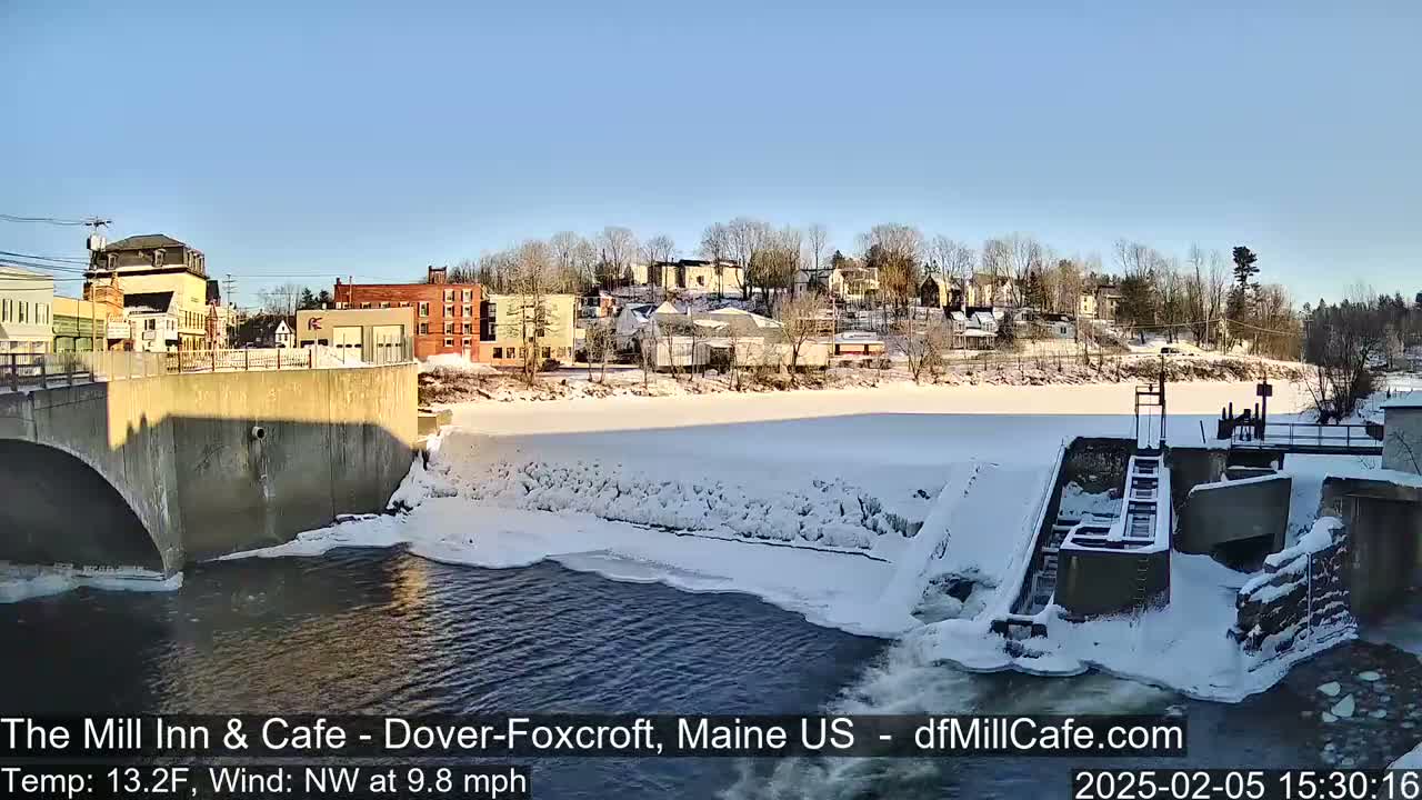 A snow-covered town is viewed from a bridge over a partially frozen river with a dam and lock system in the foreground, under a clear blue sky.