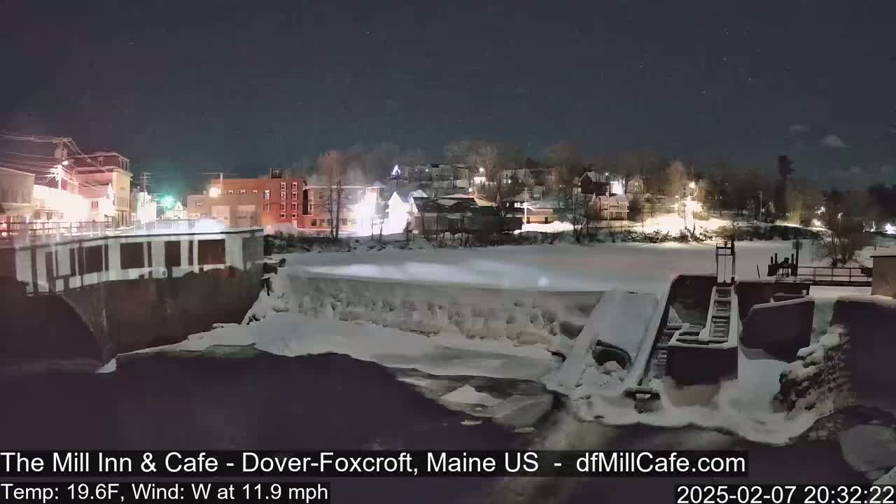 A nighttime view of a snow-covered dam and river, with a town illuminated in the background under a clear sky, and a west wind of 11.9 mph at 19.6°F.